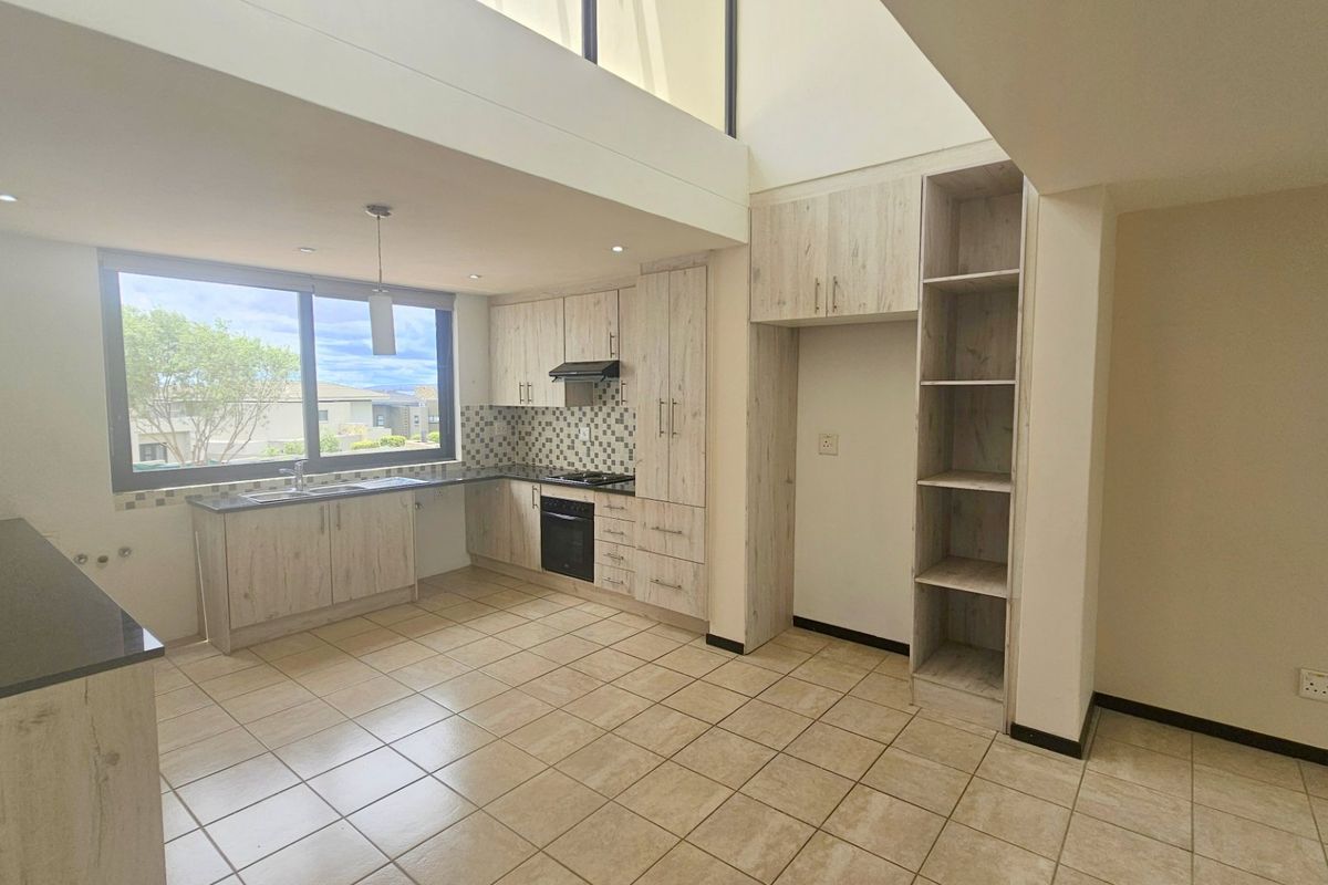 Modern kitchen featuring light wood-grain cabinetry, dark countertops, built-in oven, hob, extractor fan, and tiled flooring in an open-plan layout with double-volume ceiling.