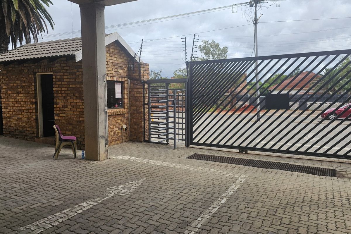 Commercial property entrance featuring a brick security guard house, paved driveway, and a large sliding metal security gate with electric fencing.