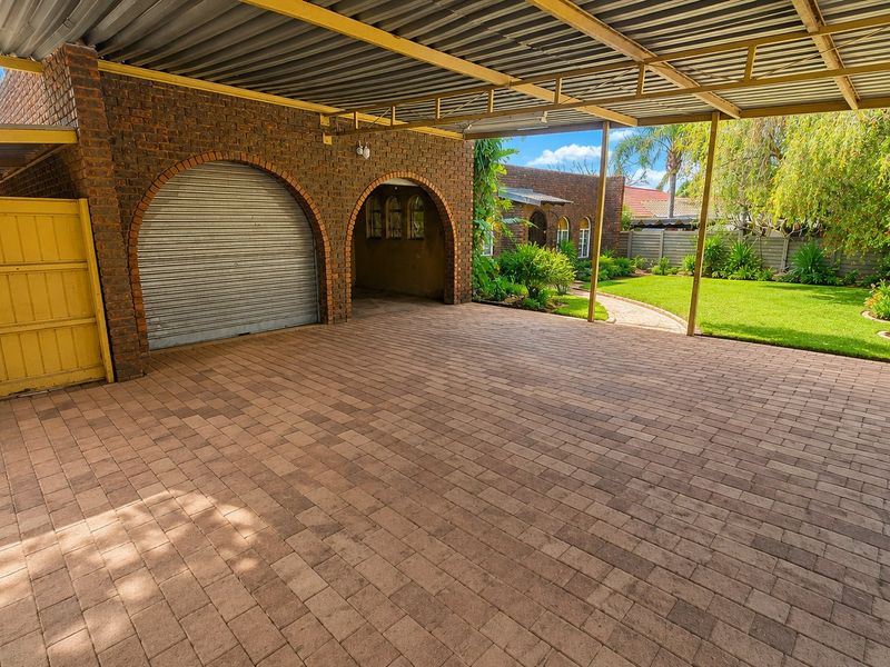 A spacious covered carport area featuring brick construction, arched architectural details, and a paved driveway leading to a secure roller door garage.