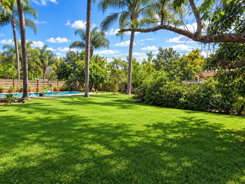 A spacious, well-maintained garden featuring a lush green lawn, mature trees, and a swimming pool enclosed by a boundary wall.