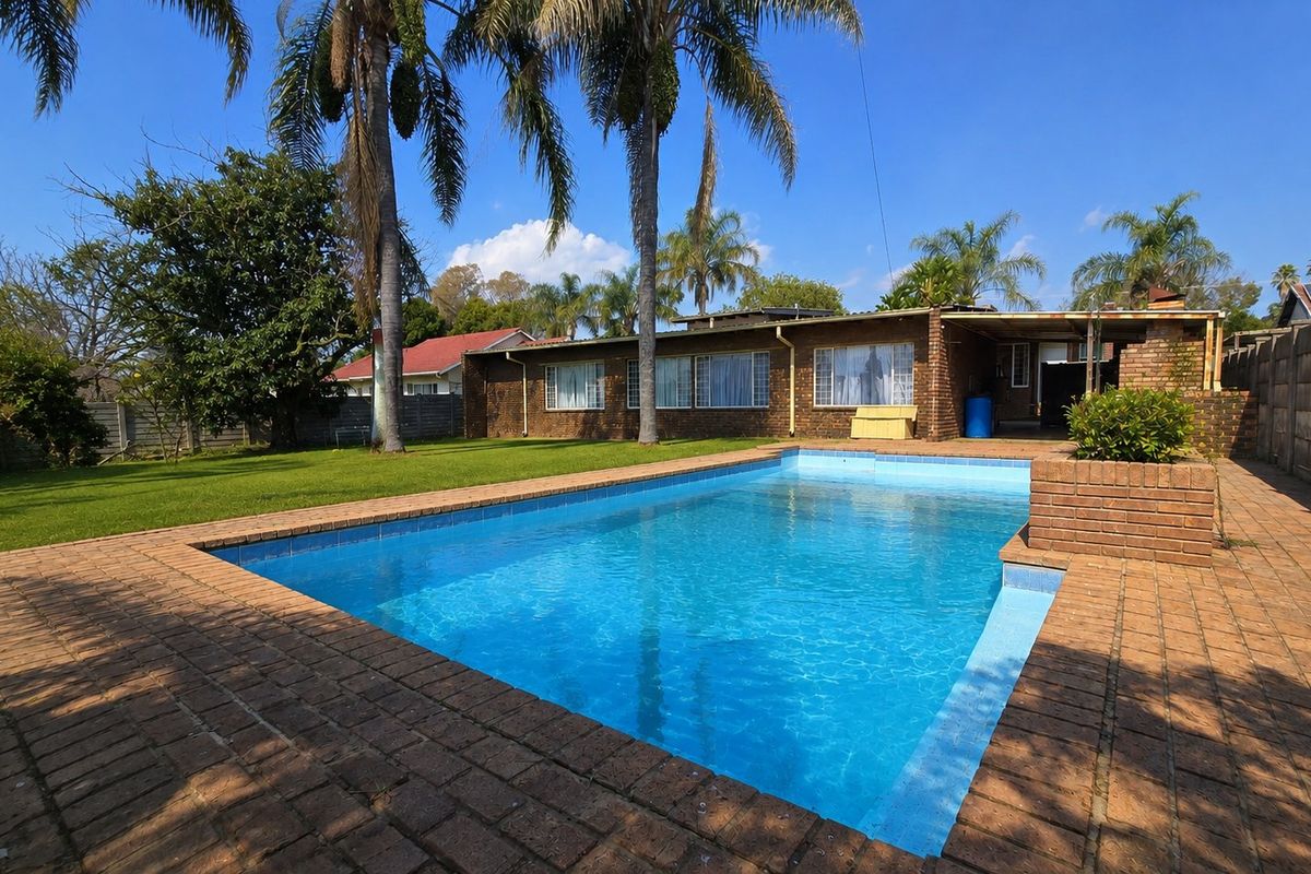 A spacious backyard featuring a swimming pool with brick paving, a covered patio area, and established palm trees set against a brick house exterior.