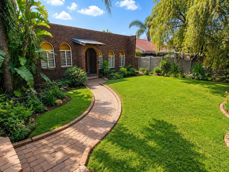 A well-maintained rear garden featuring a brick-paved pathway leading to a charming brick facade with arched windows and lush landscaping.
