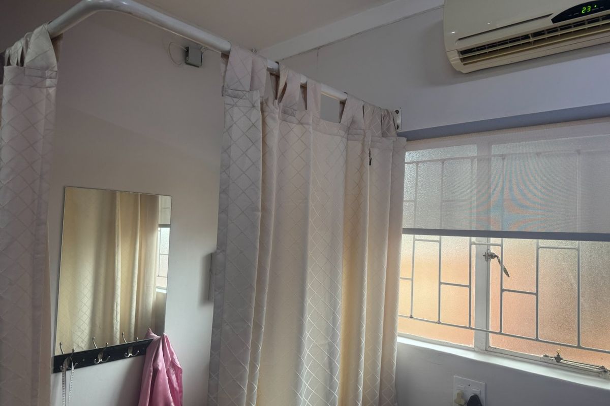 A bedroom interior featuring a wall-mounted air conditioning unit, security bars on the window, and a curtain rail setup.
