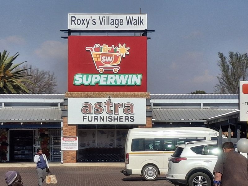 Front exterior view of Roxy's Village Walk commercial center featuring prominent signage for Superwin and Astra Furnishers with an adjacent paved parking area.