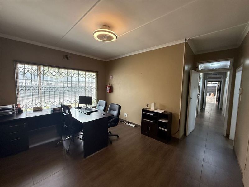 A functional office space featuring tiled flooring, security bars on the window, and direct access to the hallway.