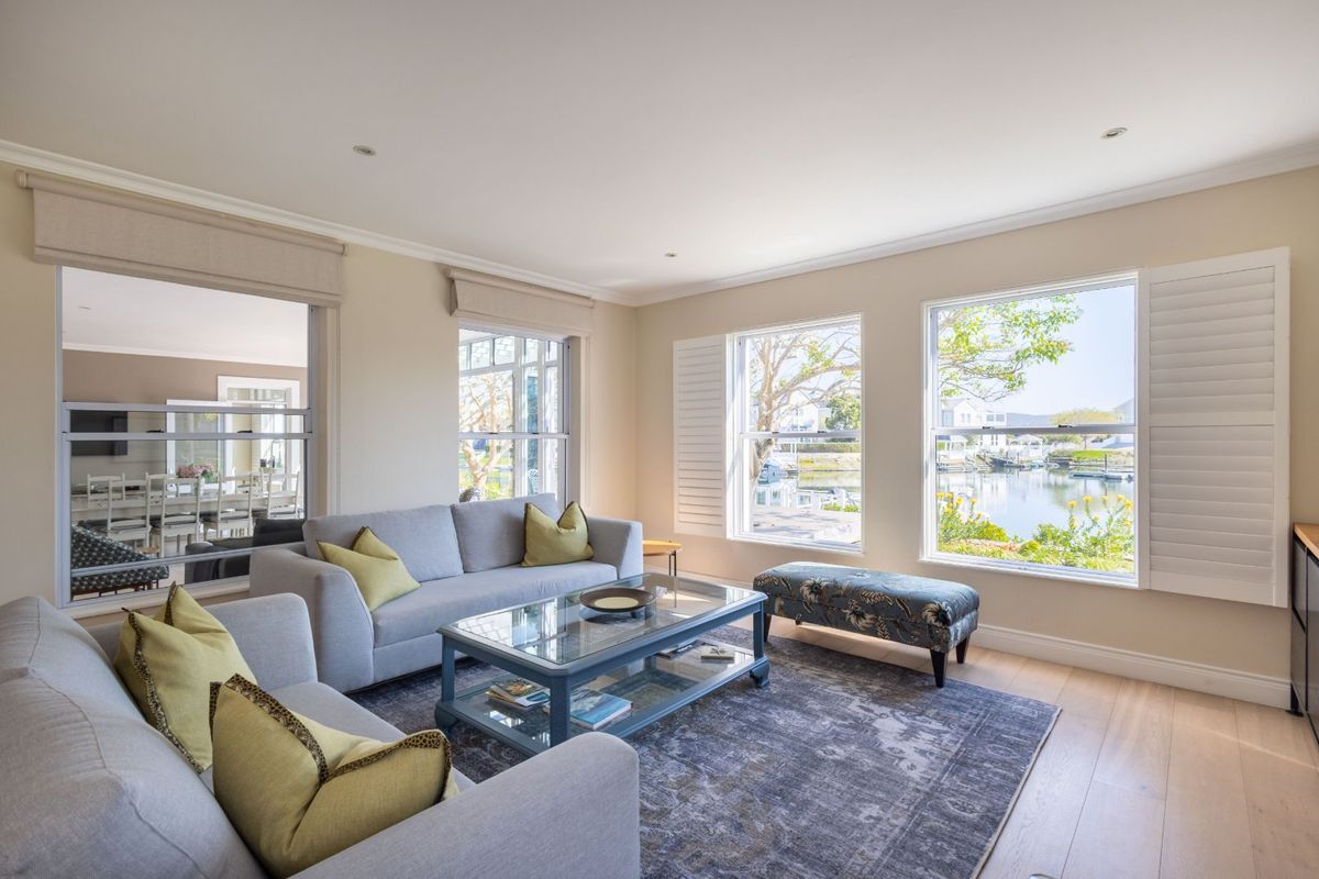 Bright and airy living room featuring timber flooring, plantation shutters, and large windows offering scenic water views.