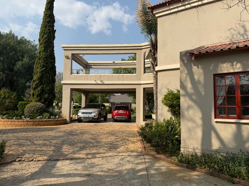 Front elevation of a multi-story house featuring a paved driveway, covered parking area, and an upper-level balcony with a glass balustrade.