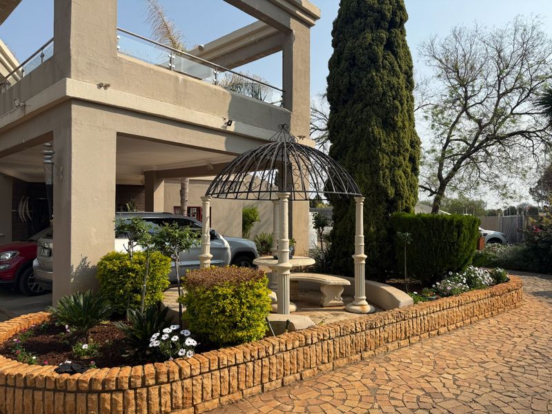 Front exterior view featuring a paved driveway, raised brick garden beds, a covered patio with glass balustrades, and a decorative wrought iron gazebo.