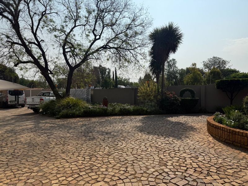 A spacious front driveway featuring patterned cobblestone paving, established trees, and a secure perimeter wall with landscaped garden beds.