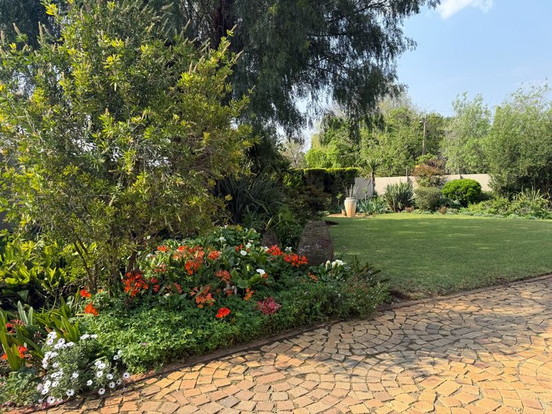 A well-maintained garden featuring a lush green lawn, mature trees, vibrant flower beds, and decorative cobblestone paving.