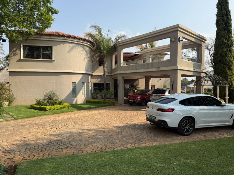 A spacious double-story residence featuring a large cobblestone driveway, covered patio, upper-level balcony, and well-maintained landscaped garden.