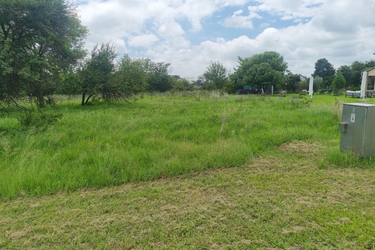 A view of a vacant residential plot featuring established trees, grass lawn, and a utility connection point.