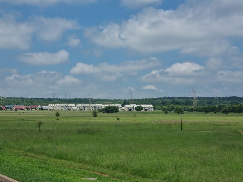 A wide-angle view of an expansive vacant residential plot featuring open grassland with distant housing developments and power lines visible on the horizon.