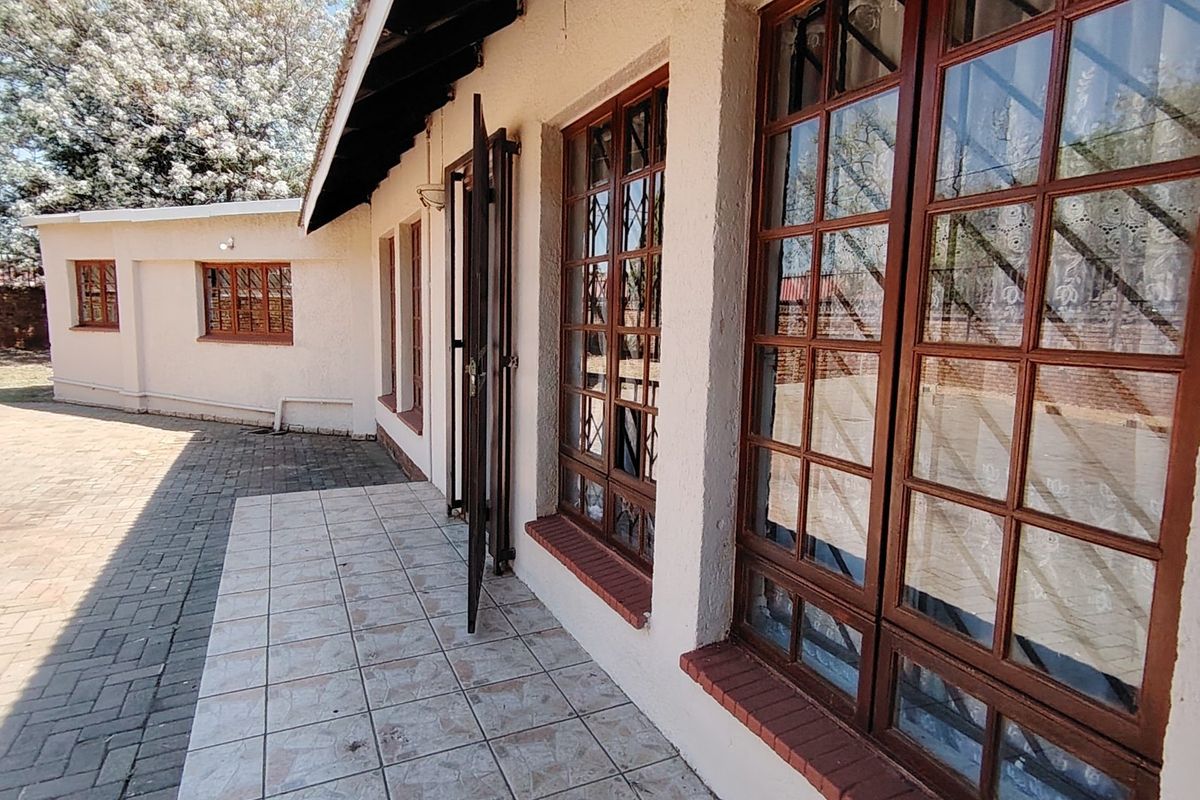 A rear exterior view featuring a paved courtyard, tiled patio area, and traditional wooden-framed windows with security bars.