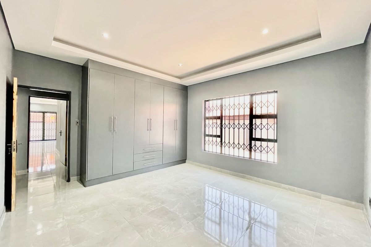 A bright, modern bedroom featuring neutral grey walls, polished tiled flooring, extensive built-in cabinetry, and a recessed ceiling with integrated downlights.