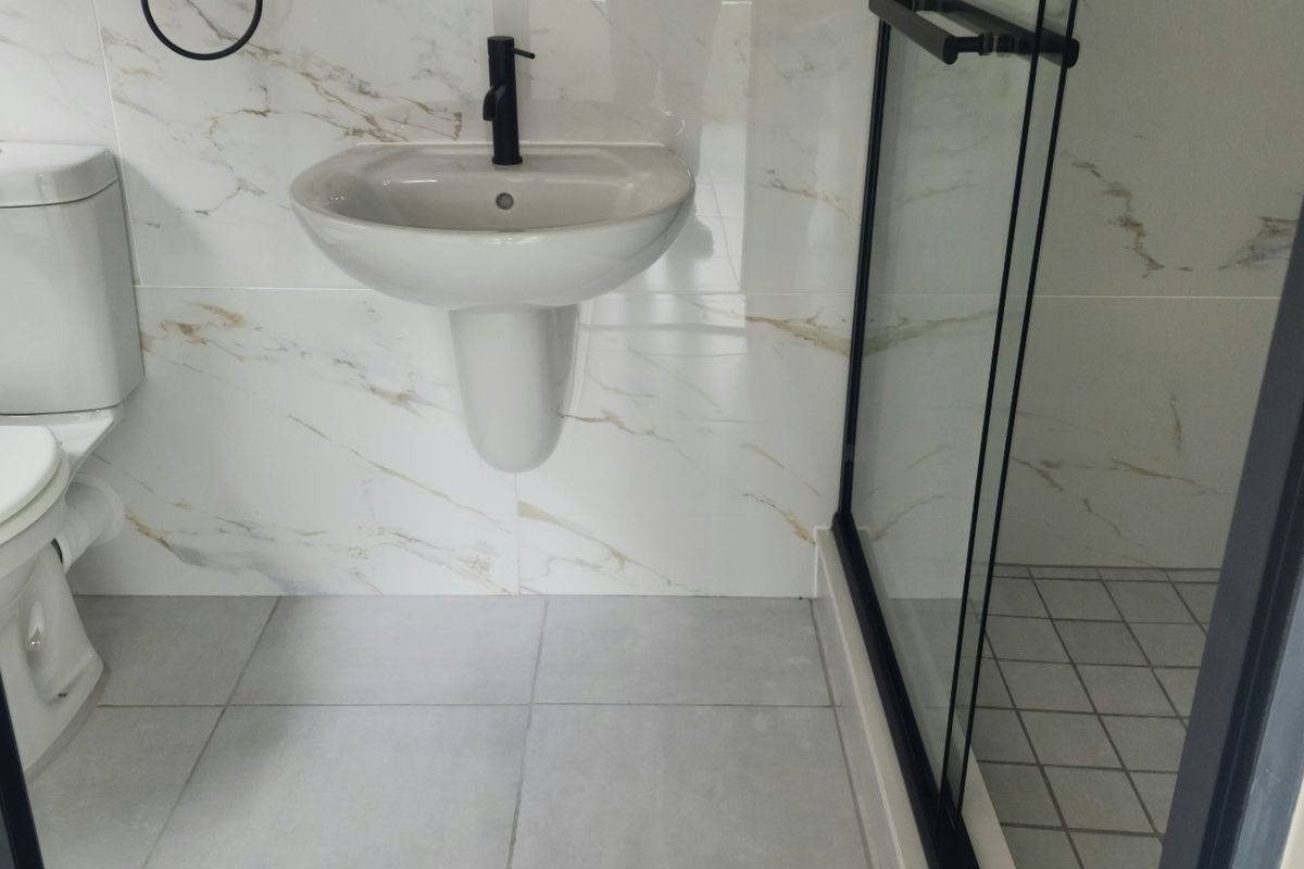 A modern, well-maintained bathroom featuring marble-effect wall tiling, a pedestal washbasin with black tapware, and a glass-enclosed shower.