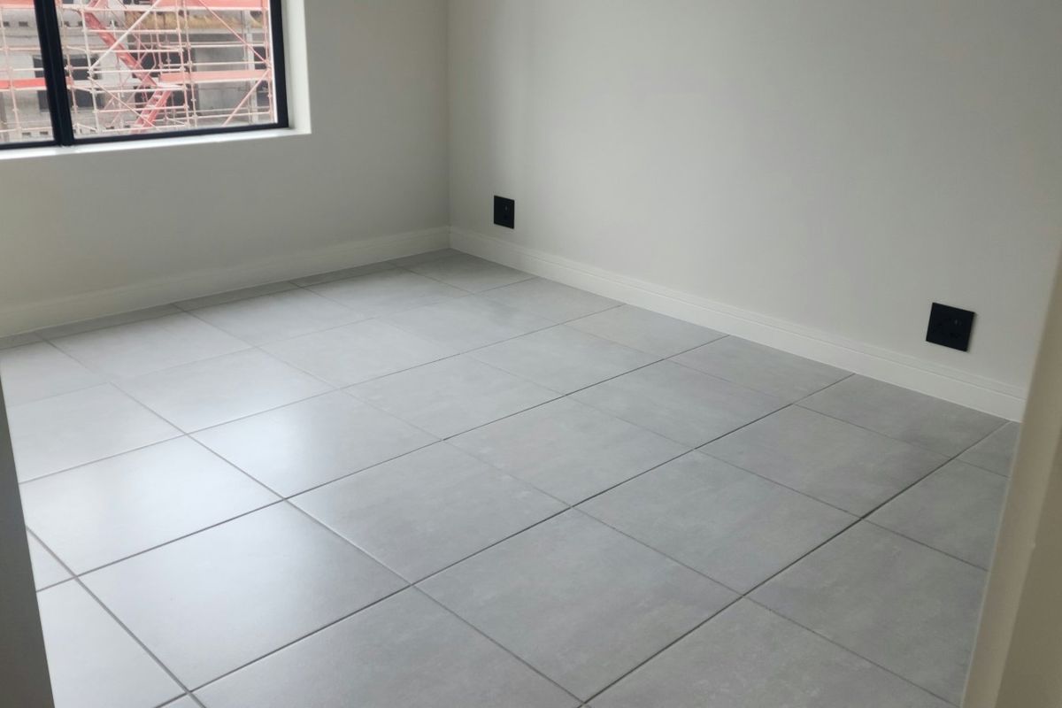 A clean, empty bedroom featuring modern grey floor tiles, white walls, and a large window with an aluminium frame.