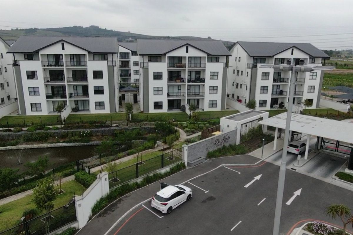 Aerial view of a modern apartment complex featuring landscaped gardens, a water feature, and a secure gated entrance with paved driveway.