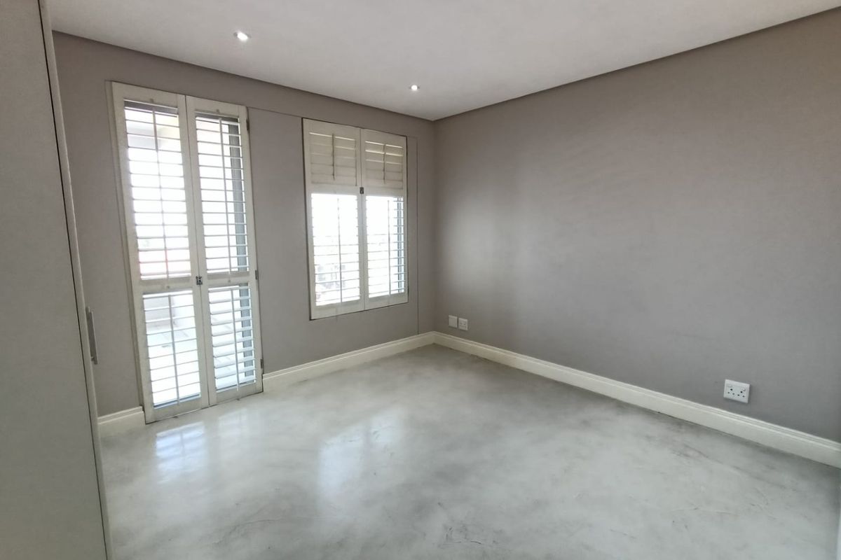 A bright, empty bedroom featuring polished concrete floors, neutral grey walls, and white plantation shutters, offering a clean and modern blank canvas.