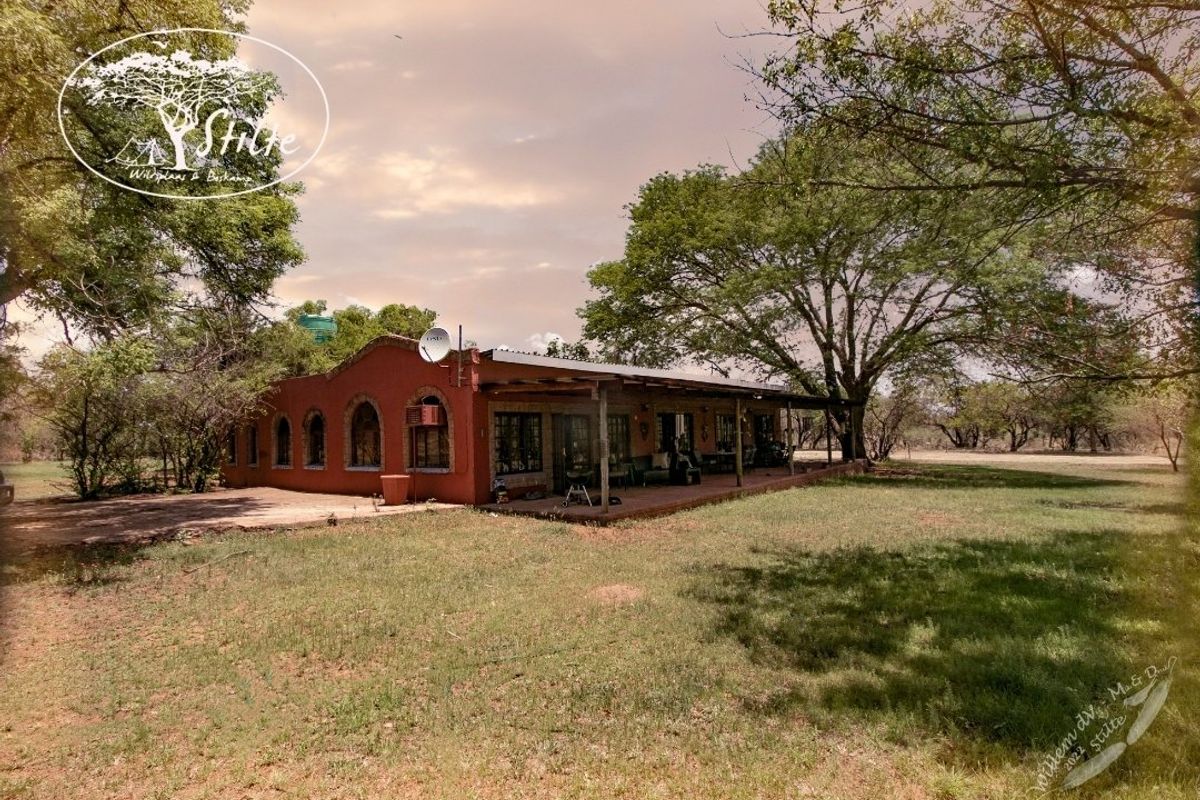 Rear view of a rustic farm dwelling featuring a covered patio, arched windows, and established surrounding trees.