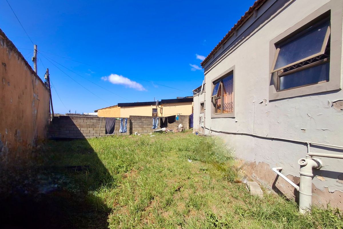 Rear exterior view of the property showing a grassed yard, boundary walls, and the side elevation of the house with visible paint deterioration.