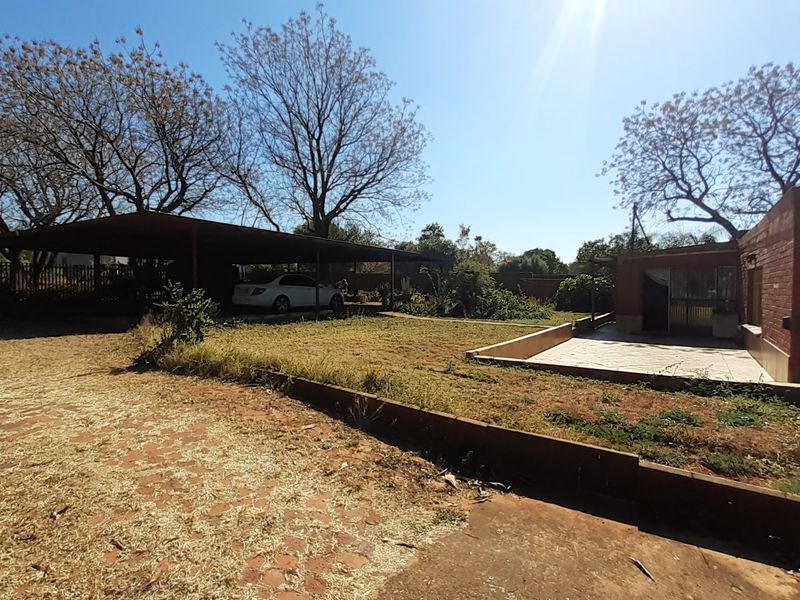 Rear exterior view of the property featuring a large carport, paved patio area, and brick building facade set within a spacious garden.