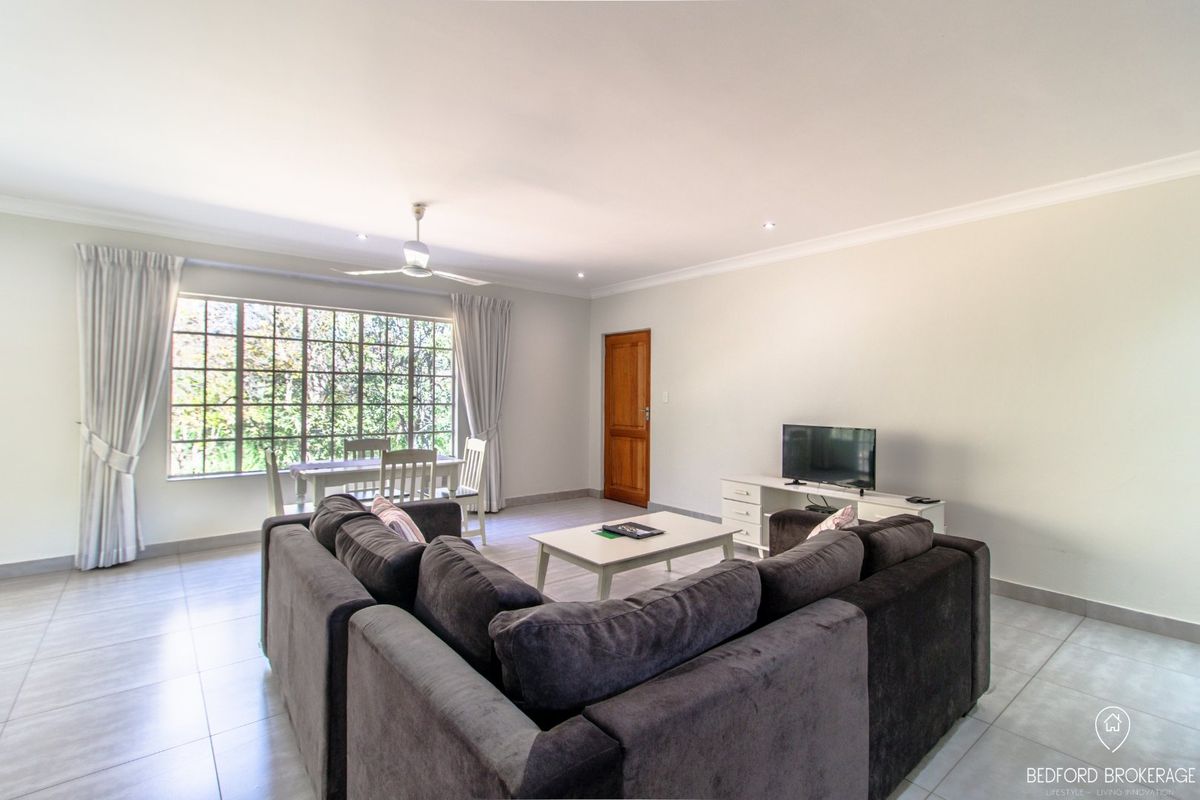 A bright and spacious living room featuring neutral tiled flooring, a ceiling fan, and large windows that provide ample natural light.