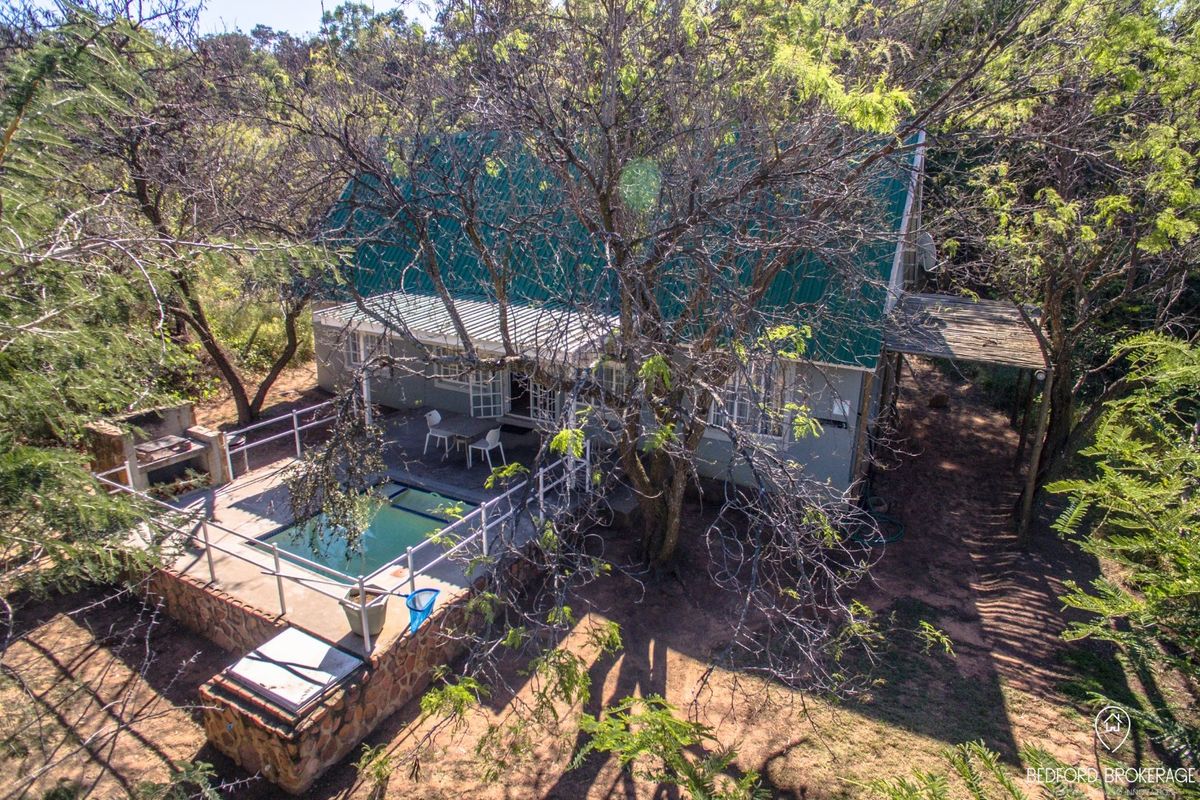 Aerial view of a farm property featuring a swimming pool, stone-walled patio area, built-in braai, and a metal-roofed dwelling surrounded by mature trees.