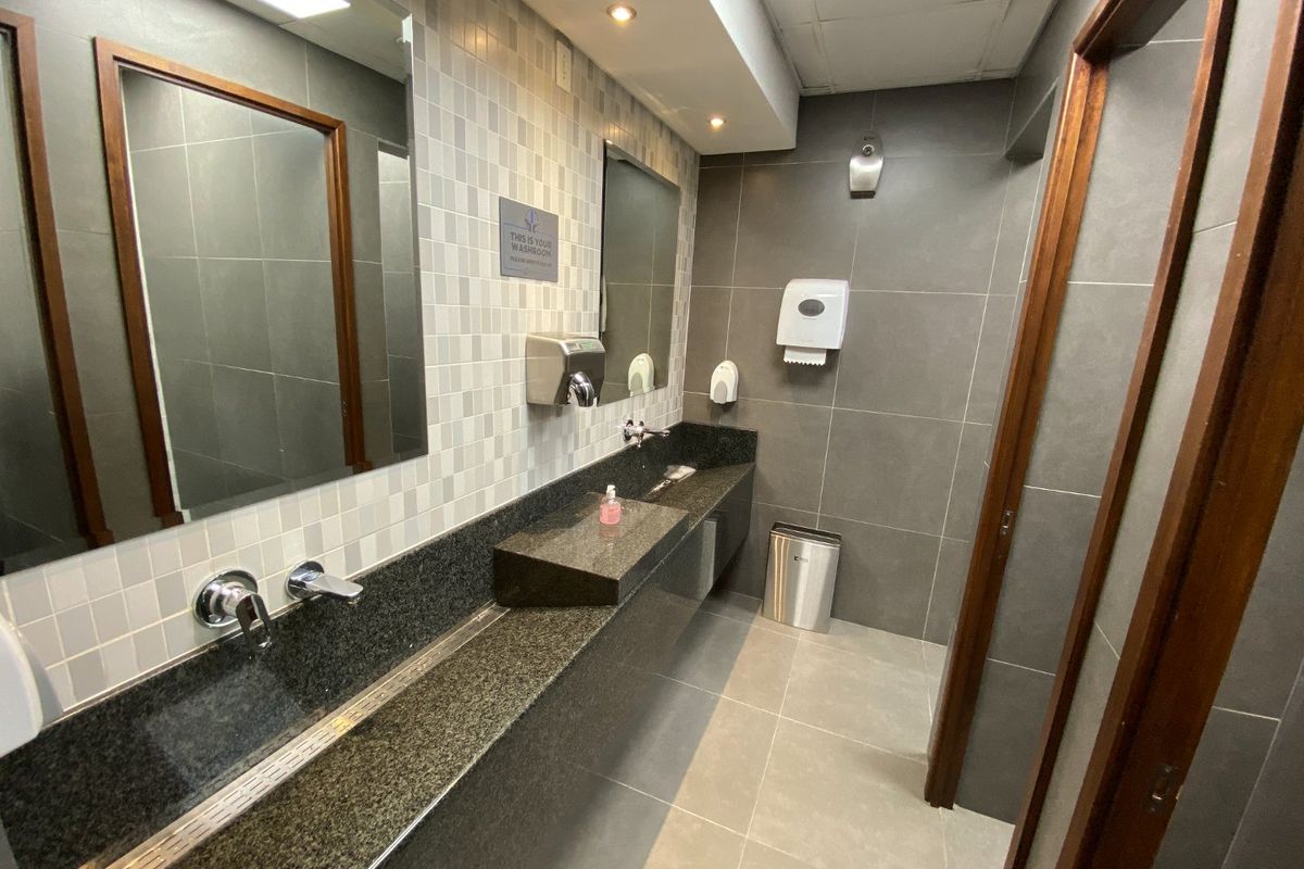 Modern commercial bathroom featuring a long granite vanity countertop with wall-mounted faucets, tiled walls, and recessed ceiling lighting.