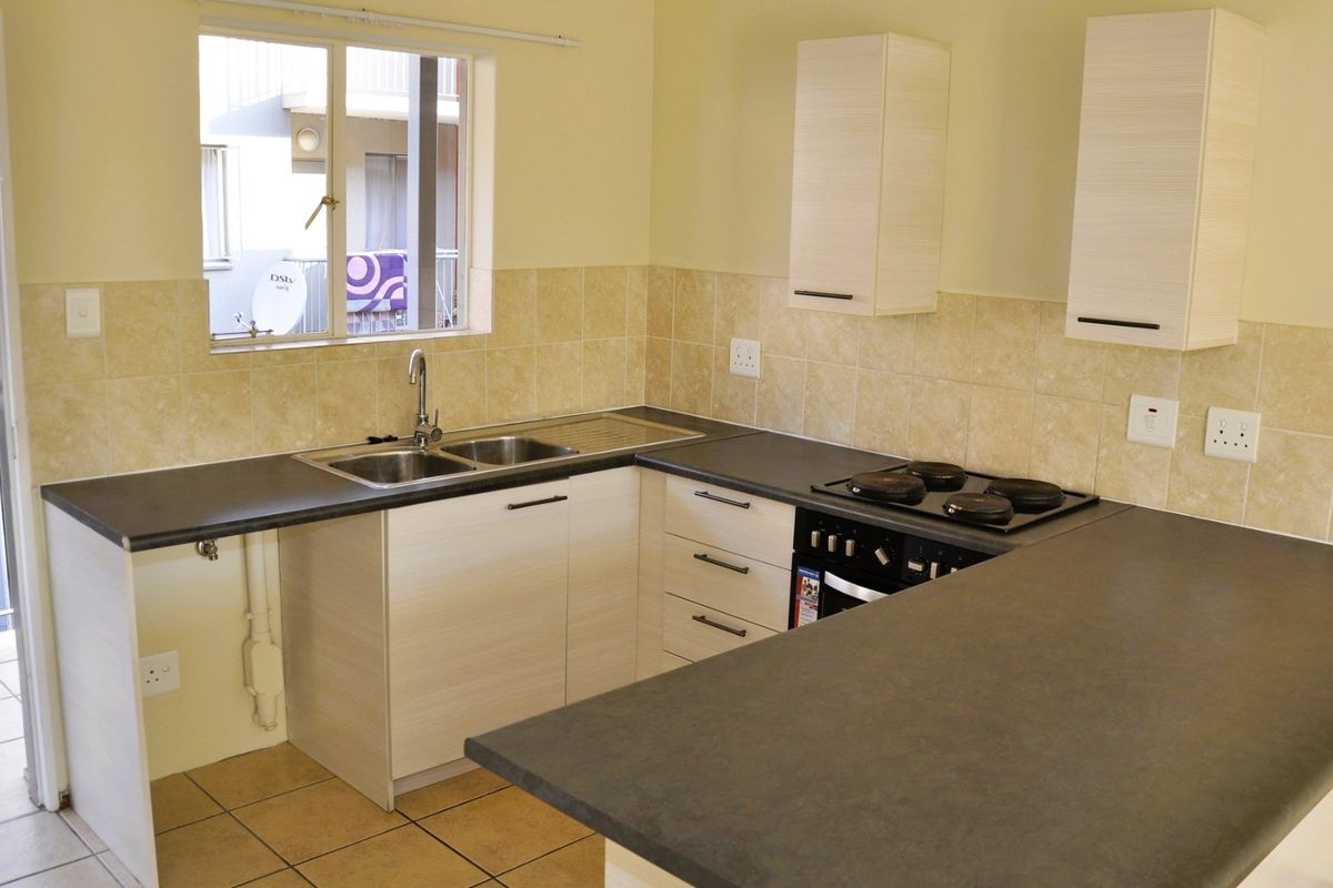 A clean, functional kitchen featuring light wood-look cabinetry, dark laminate countertops, a built-in oven with electric hob, and neutral tiled splashbacks.