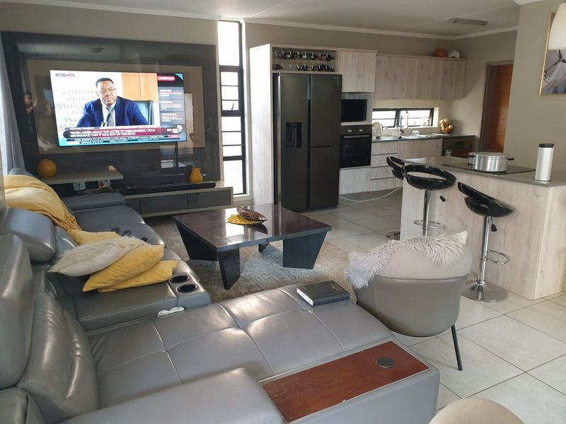 Modern open-plan living area featuring a contemporary kitchen with island, built-in appliances, and a dedicated media wall, finished with neutral tiled flooring.