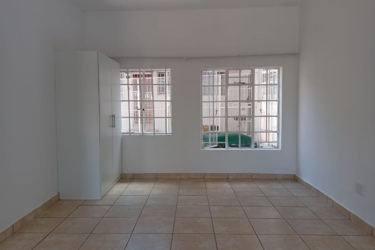 A bright, empty bedroom featuring neutral tiled flooring, a built-in wardrobe, and windows fitted with security bars.