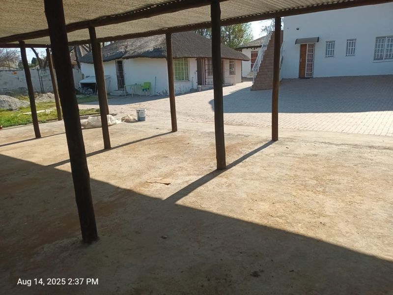 A spacious outdoor courtyard featuring a covered patio area with timber supports, paved ground, and access to an external staircase.