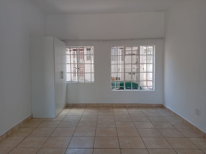 A bright, empty bedroom featuring neutral tiled flooring, a built-in wardrobe, and windows fitted with security bars.