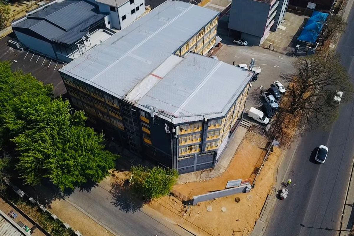 Aerial view of a multi-story commercial building featuring a metal roof, paved parking facilities, and perimeter fencing situated along a main road.