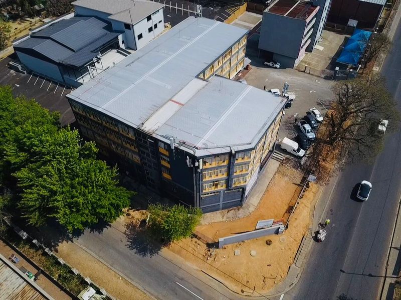 Aerial view of a multi-story commercial building featuring a metal roof, paved parking facilities, and perimeter fencing situated along a main road.