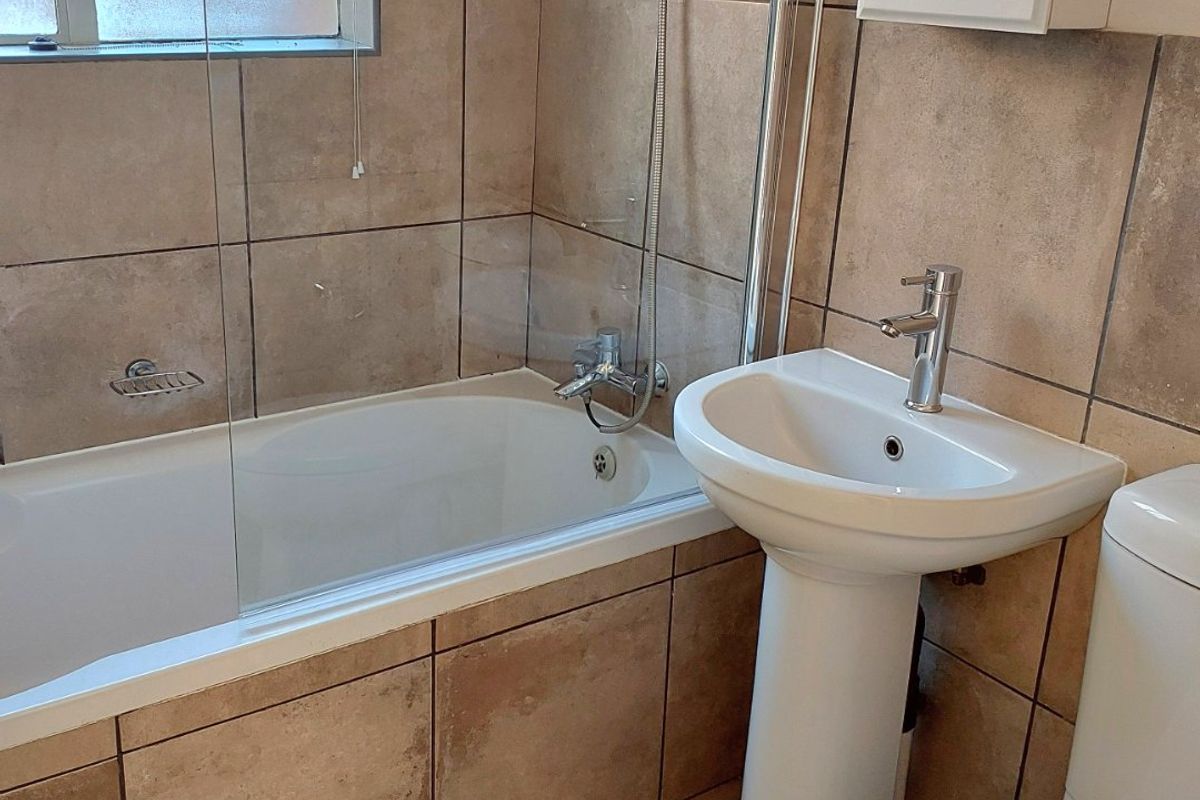 A functional bathroom featuring a bathtub with a shower attachment, glass screen, pedestal basin, and toilet, finished with neutral wall and floor tiling.