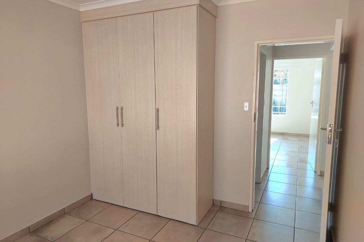 A clean, empty bedroom featuring light wood-finish built-in wardrobes, neutral wall paint, and tiled flooring, with a view into the adjacent hallway.
