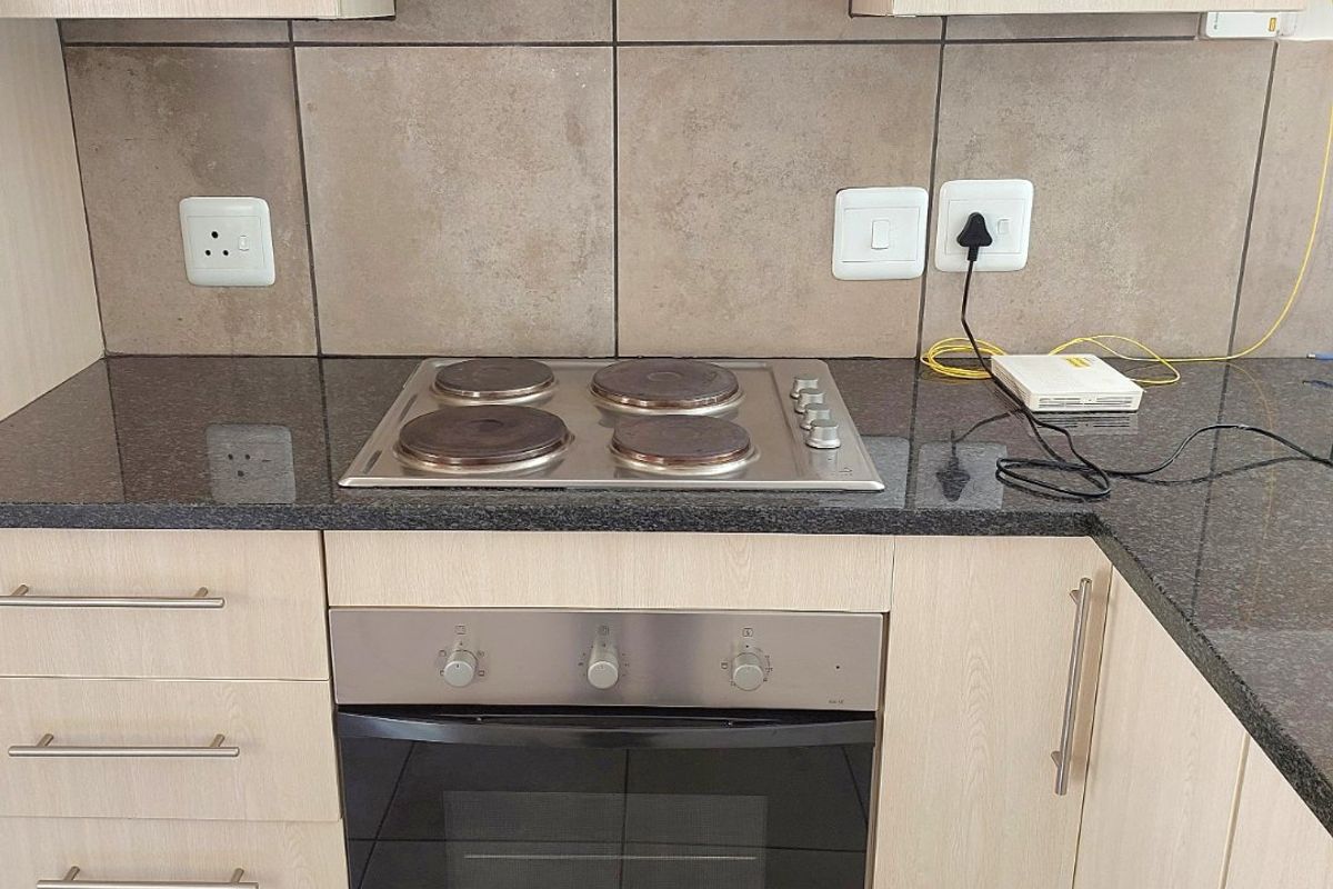 A clean, functional kitchen featuring light wood-finish cabinetry, granite countertops, a built-in oven, electric hob, and a stainless steel extractor fan.