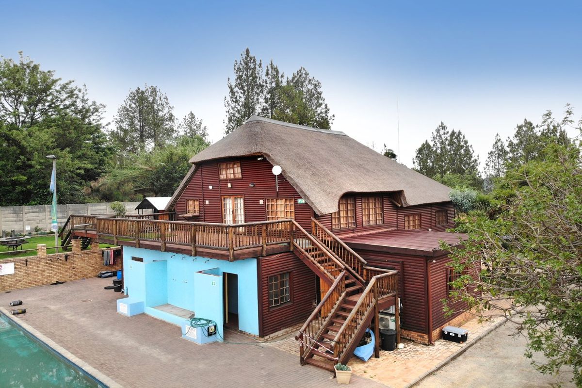 Rear view of a timber log building featuring a thatched roof, a raised wooden deck with staircase, and an adjacent paved pool area.