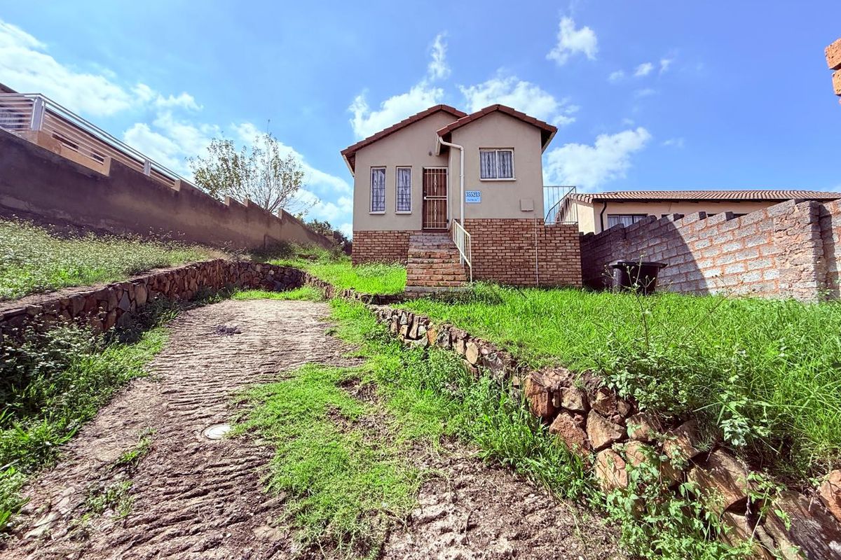 Front view of a detached house featuring a brick base, tiled roof, and a sloped garden with stone retaining walls and a driveway.