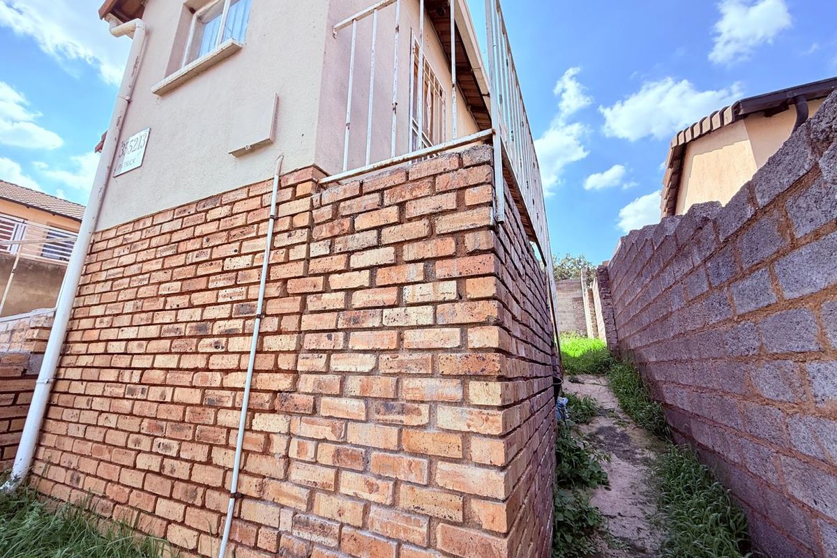 Side exterior view of the property featuring exposed brickwork, a metal balcony railing, and a narrow pathway between boundary walls.