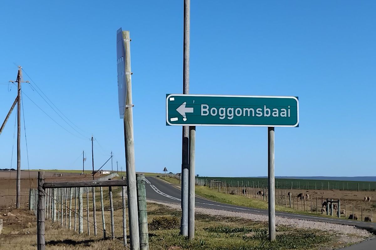 A view of a rural road and directional signage indicating the way to Boggomsbaai, surrounded by open fields and wire fencing.