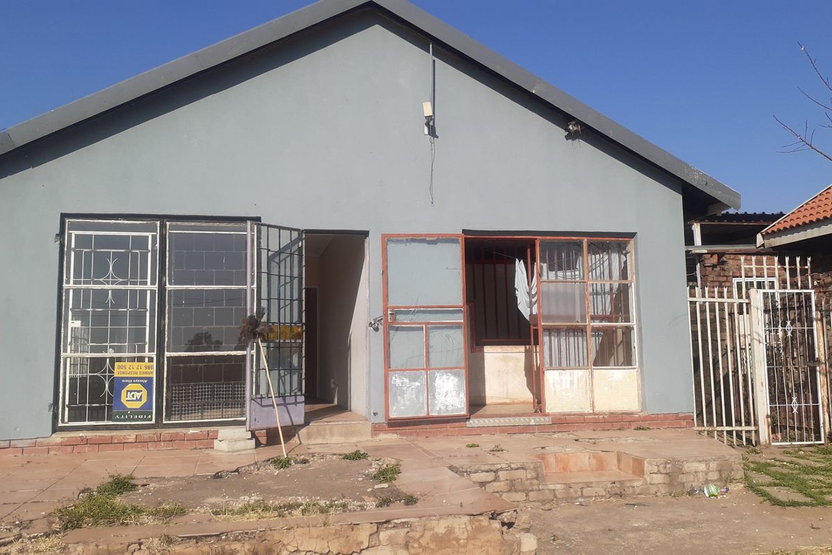 Rear exterior view of the house featuring a paved patio area, security gates, and metal window frames requiring maintenance and exterior refurbishment.