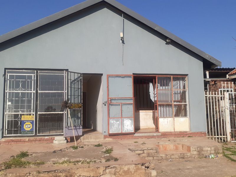 Rear exterior view of the house featuring a paved patio area, security gates, and metal window frames requiring maintenance and exterior refurbishment.
