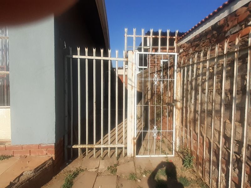 A narrow side passage featuring metal security gates, brick boundary walls, and palisade fencing, showing signs of wear and weathering.