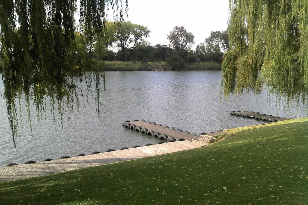 A scenic view of a calm riverfront featuring a wooden jetty and well-maintained grassy banks framed by weeping willow trees.