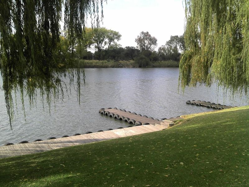 A scenic view of a calm riverfront featuring a wooden jetty and well-maintained grassy banks framed by weeping willow trees.