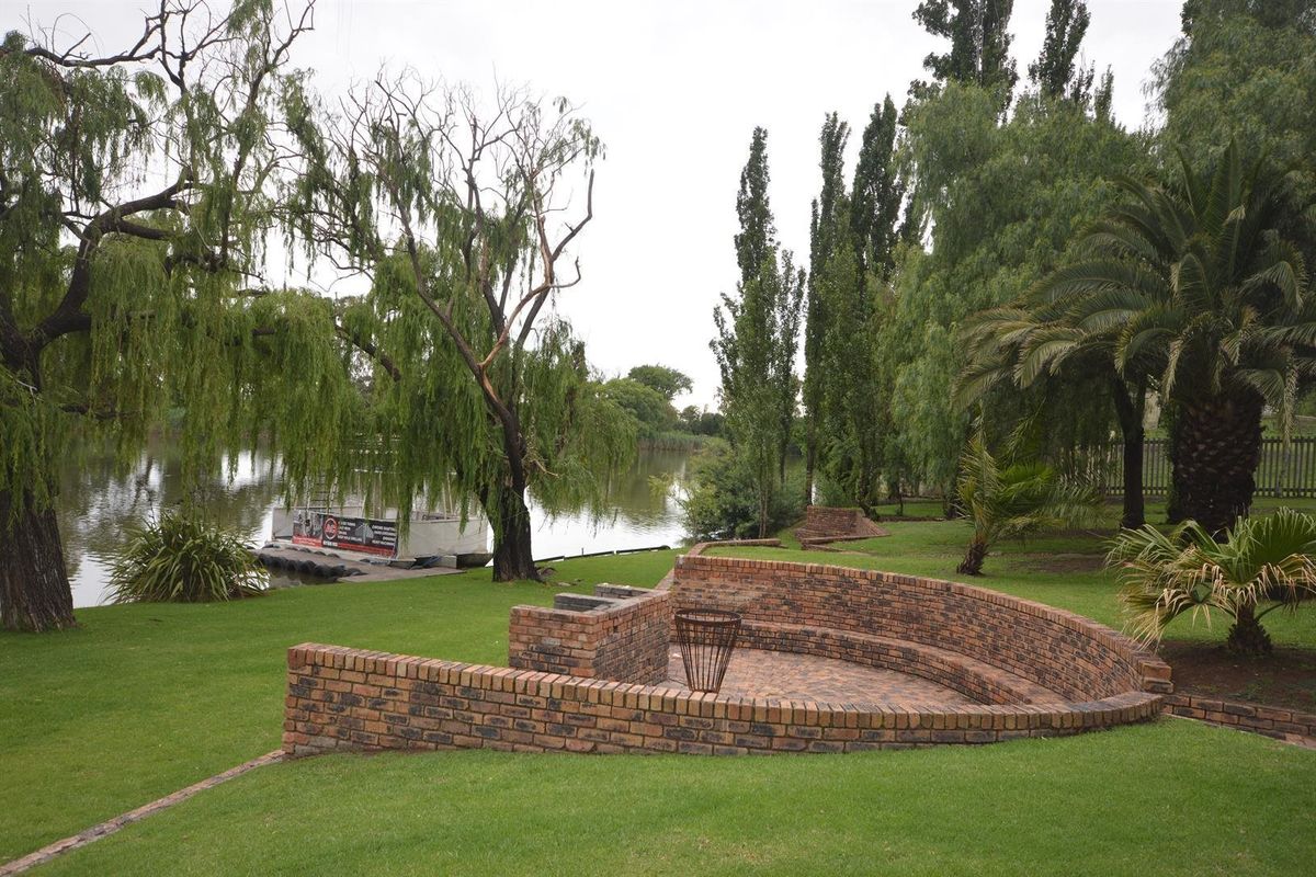 A well-maintained garden area featuring a curved brick seating wall, built-in braai, and paved patio overlooking the water.