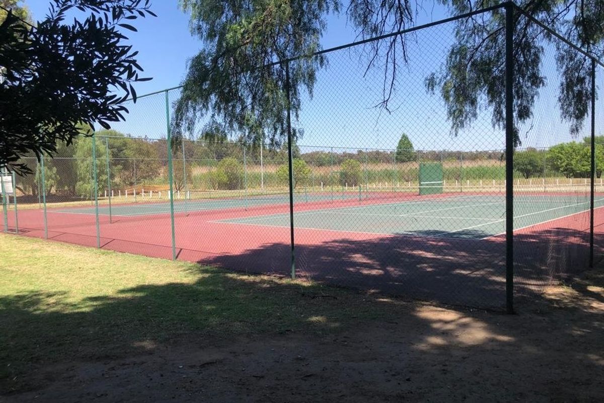 Outdoor tennis court facility featuring a hard surface and perimeter chain-link fencing, situated in a landscaped estate setting.